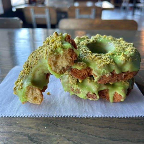 Two pistachio donuts with green glaze and crushed pistachios, one broken in half, on white paper.