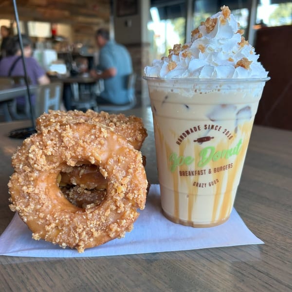 Joe Donut's iced coffee with whipped cream and two glazed donuts with crumbles on a table.