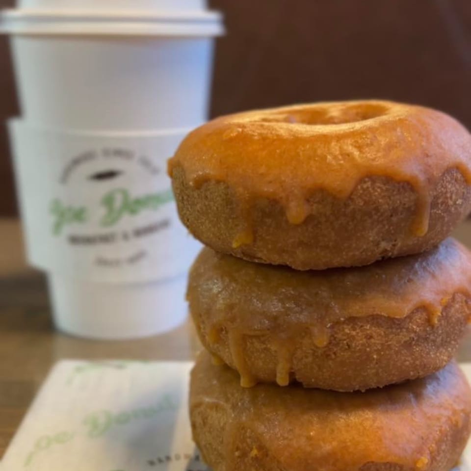 Three glazed Joe Donut donuts stacked next to a branded coffee cup and napkin.