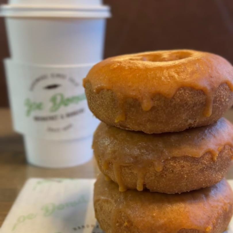Three glazed Joe Donut donuts stacked next to a branded coffee cup and napkin.