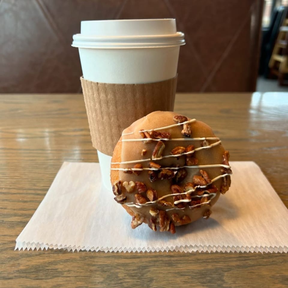 A Joe Donut coffee cup and a pecan-topped donut with white icing drizzle on a white napkin.