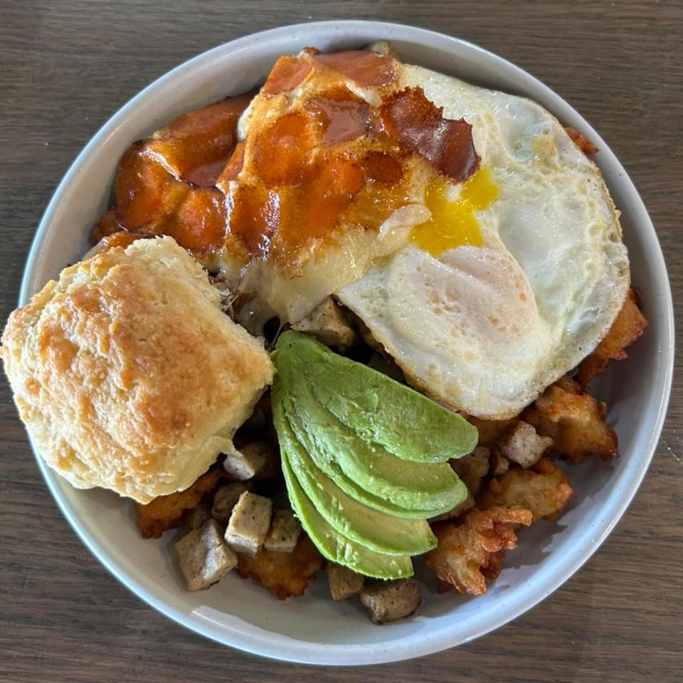 A breakfast bowl with fried eggs, avocado slices, a biscuit, and sausage gravy.