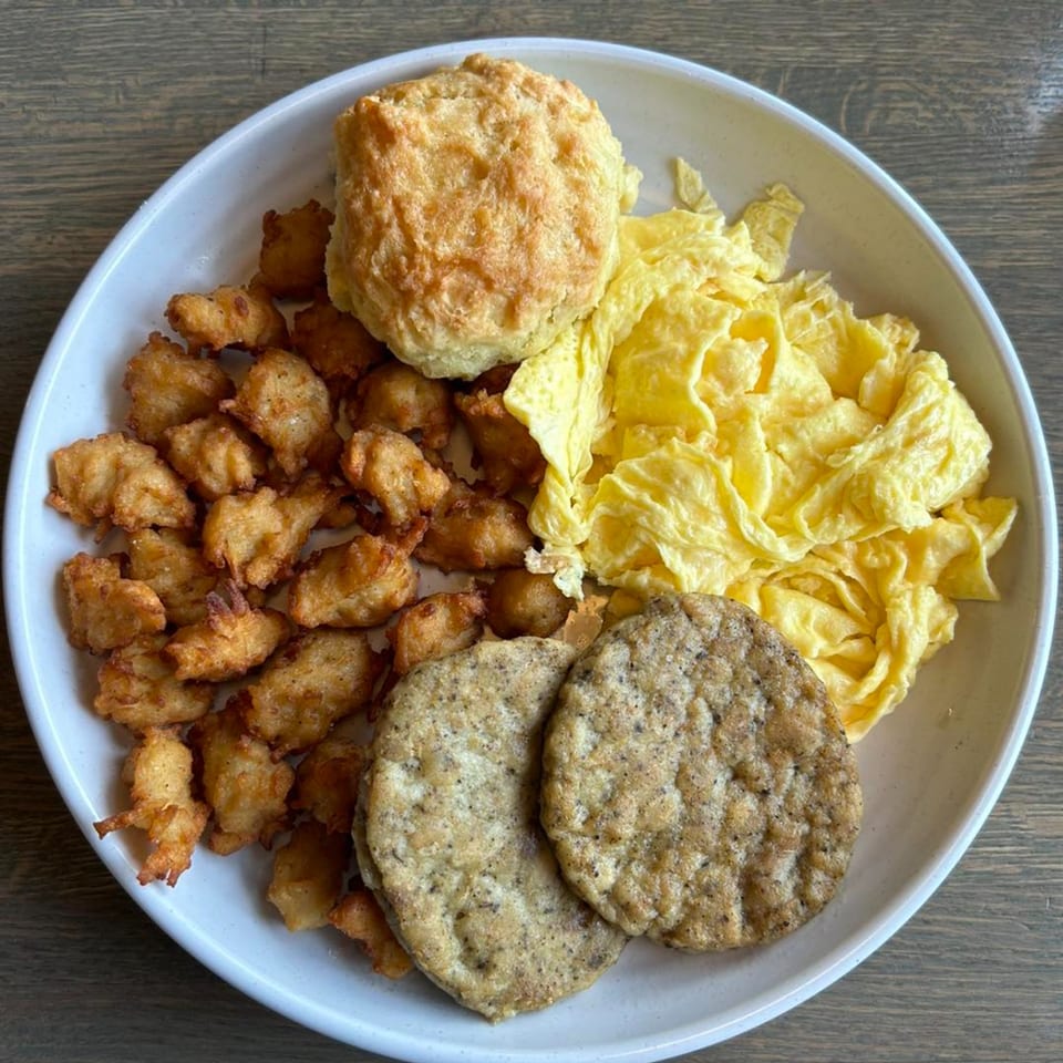 A breakfast plate with scrambled eggs, fried chicken bites, a biscuit, and two sausage patties.