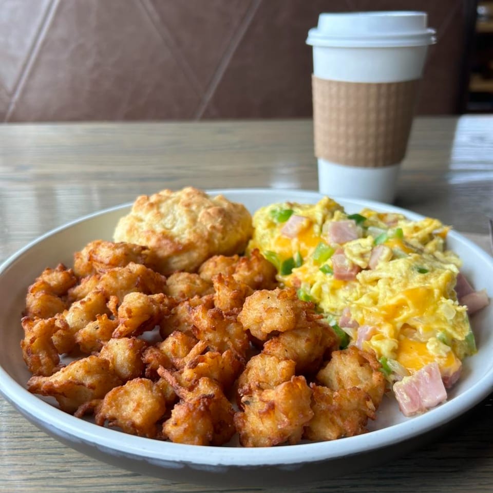 A breakfast plate with crispy tater tots, scrambled eggs with ham and peppers, a biscuit, and a coffee cup.