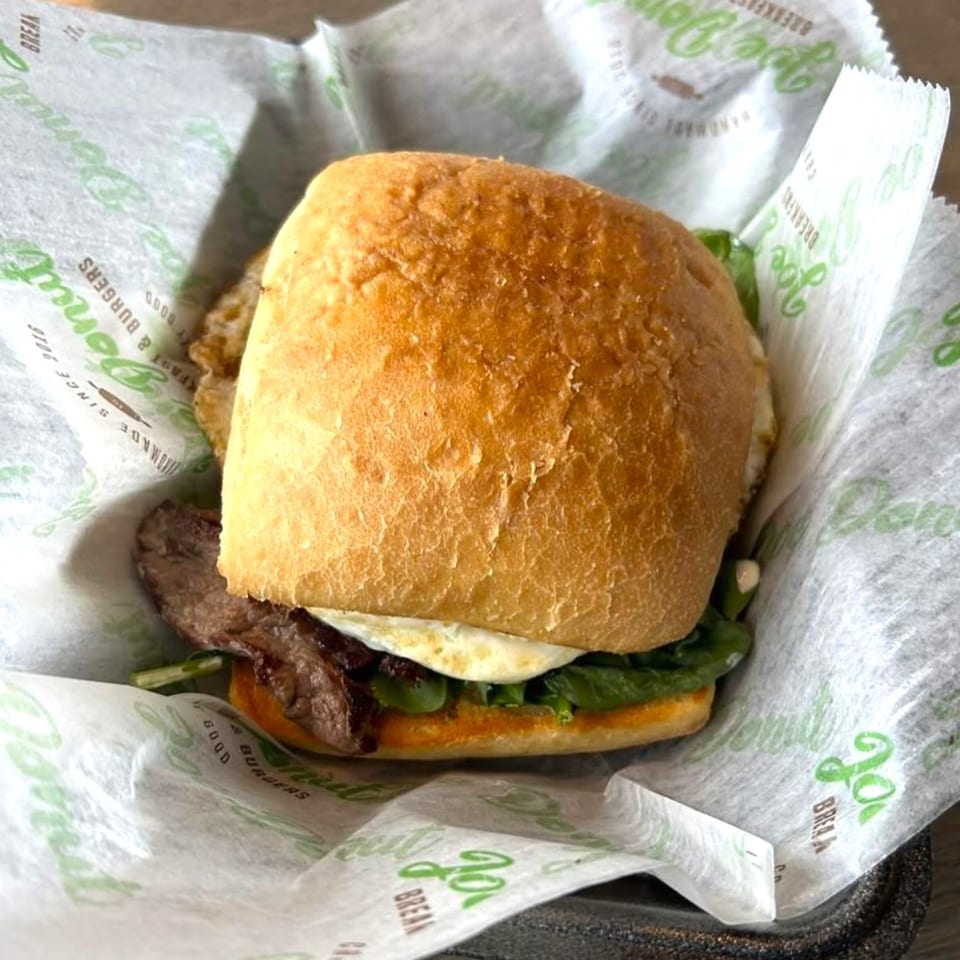 A Joe Donut breakfast sandwich with egg, meat, and greens on a bun, wrapped in branded paper.
